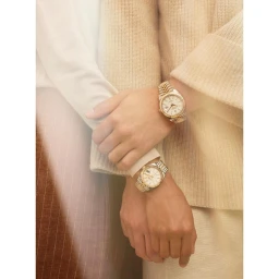 Close-up of two people wearing matching gold and silver wristwatches with white dials against a warm neutral background.