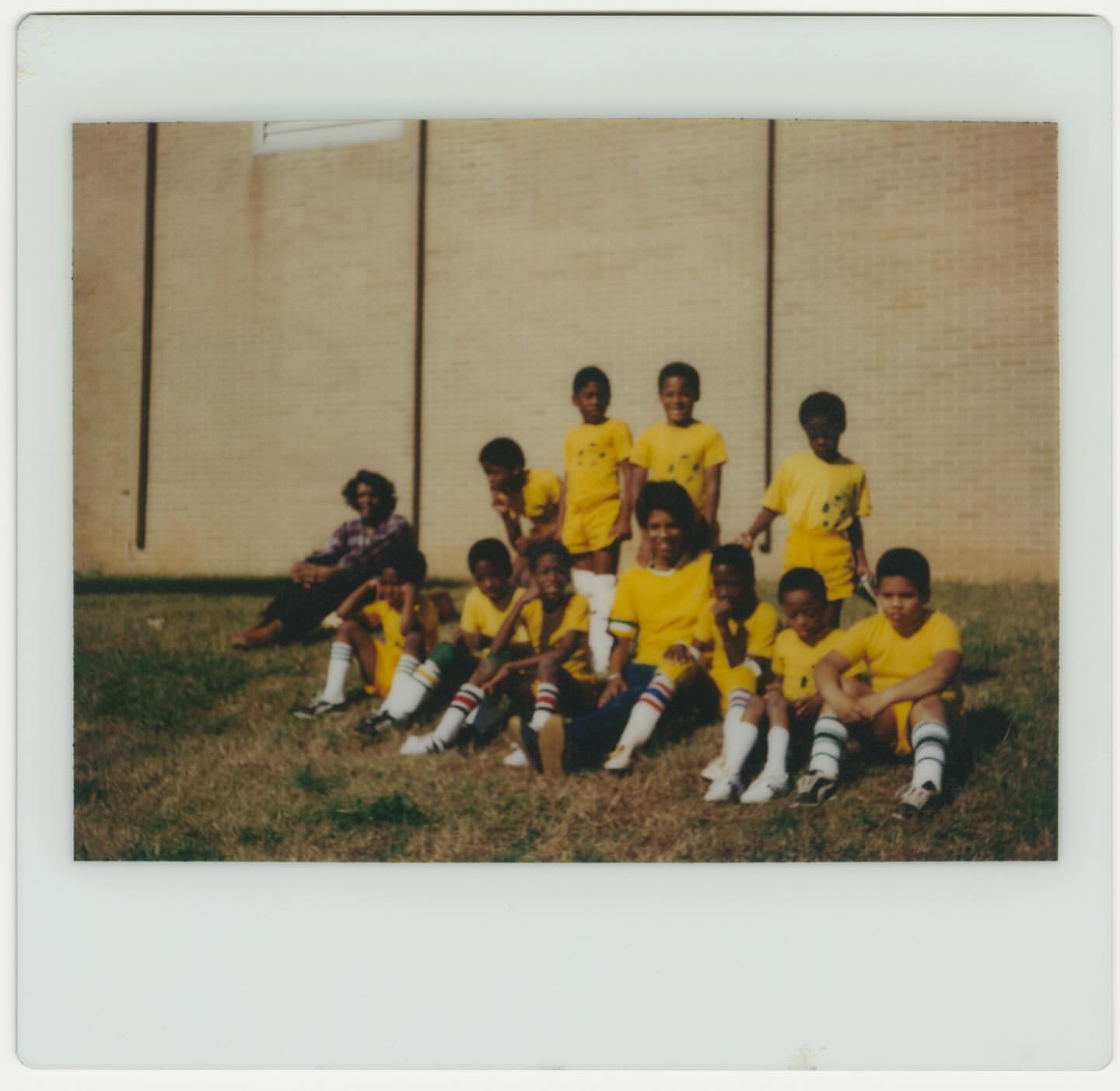A group of children's soccer team in yellow uniforms posing together on a grassy field in front of a brick building on a sunny day.