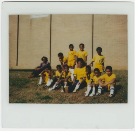 A group of children's soccer team in yellow uniforms posing together on a grassy field in front of a brick building on a sunny day.