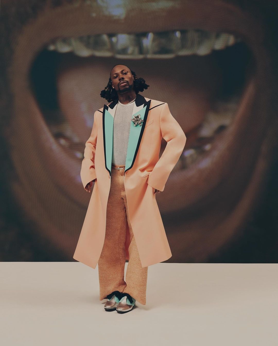 Full-length portrait of an adult male wearing a peach-colored coat and pants, posed in front of a background featuring a mouth with gold teeth.