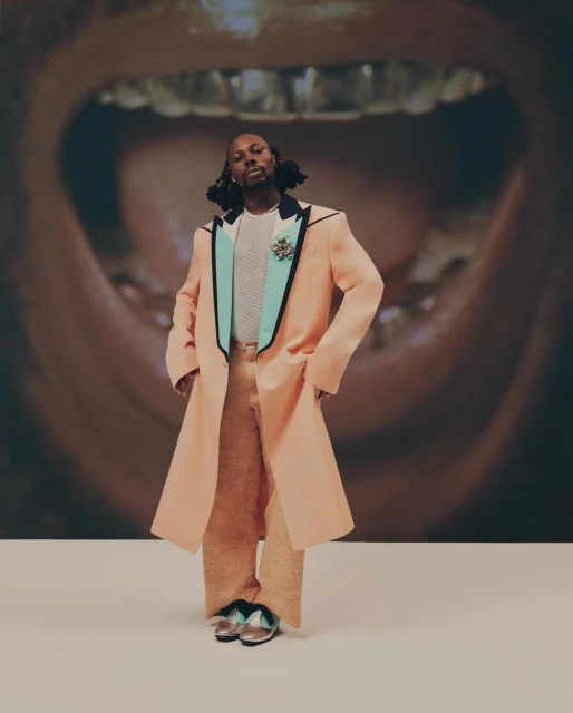 Full-length portrait of an adult male wearing a peach-colored coat and pants, posed in front of a background featuring a mouth with gold teeth.