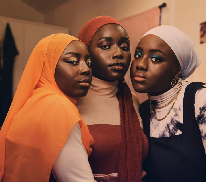 Three Black women pose indoors wearing colorful headscarves, makeup, and fashionable outfits. The shot is a medium portrait.