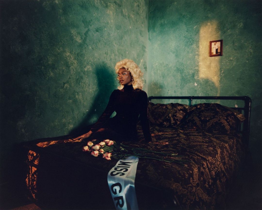 Black woman with blonde curly hair wearing a black long-sleeve top sitting on a patterned bed in a green textured room