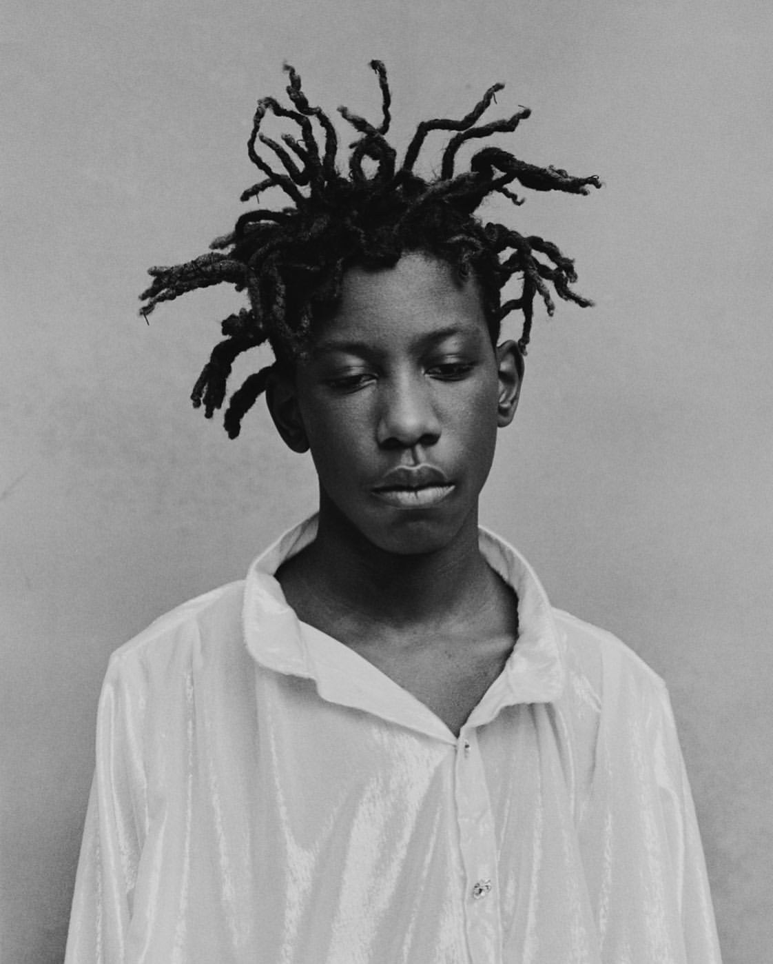 Monochrome portrait of a young person with dreadlocks, looking down, wearing a collared shirt against a solid grey backdrop.