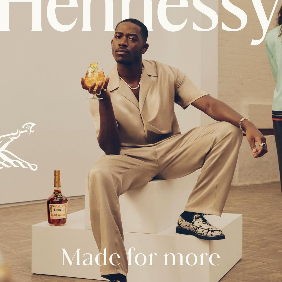 A handsome man in a tan suit sits in an art gallery, holding a cocktail, next to a Hennessy bottle in a modern advertisement.