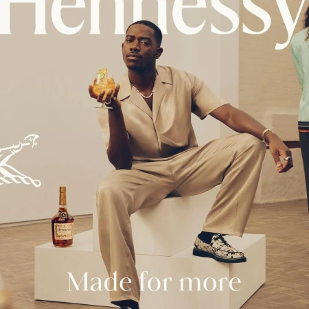A handsome man in a tan suit sits in an art gallery, holding a cocktail, next to a Hennessy bottle in a modern advertisement.