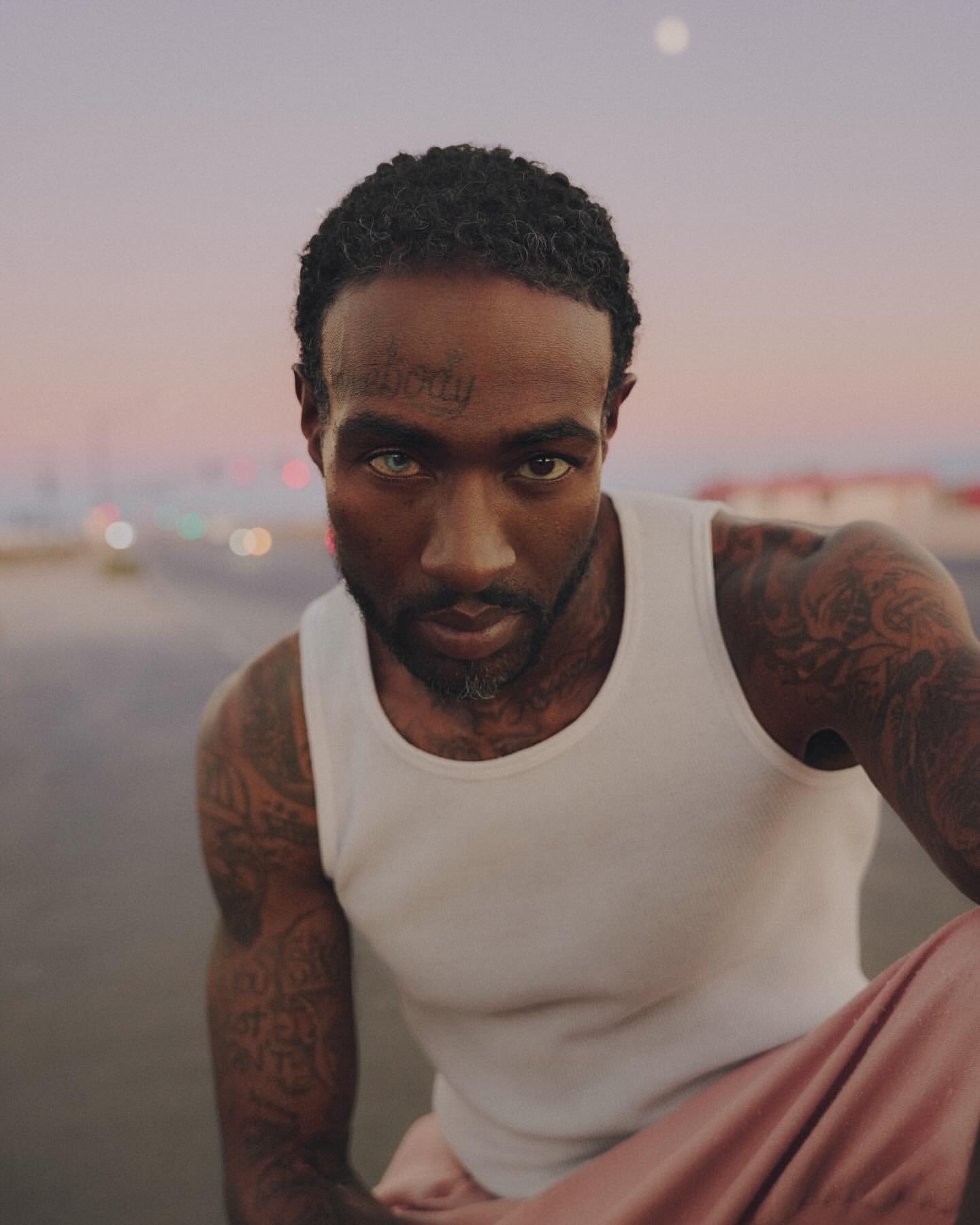 Close-up portrait of a man with tattoos wearing a white tank top, looking directly at the camera under a soft, evening sky.