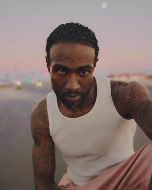 Close-up portrait of a man with tattoos wearing a white tank top, looking directly at the camera under a soft, evening sky.