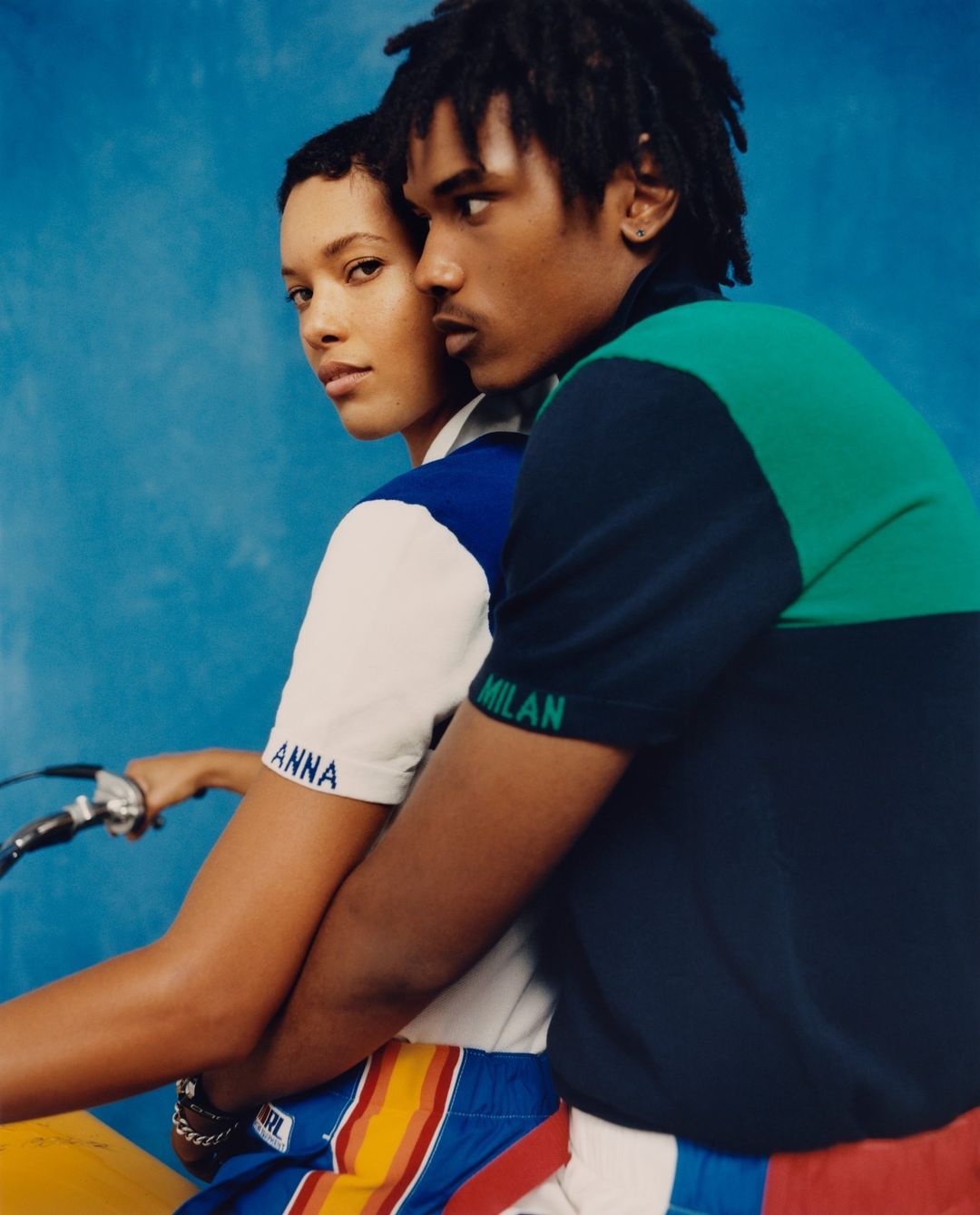 Two young adults in stylish polo shirts and shorts are captured against a textured blue backdrop, evoking a vibrant, fashion-forward aesthetic.