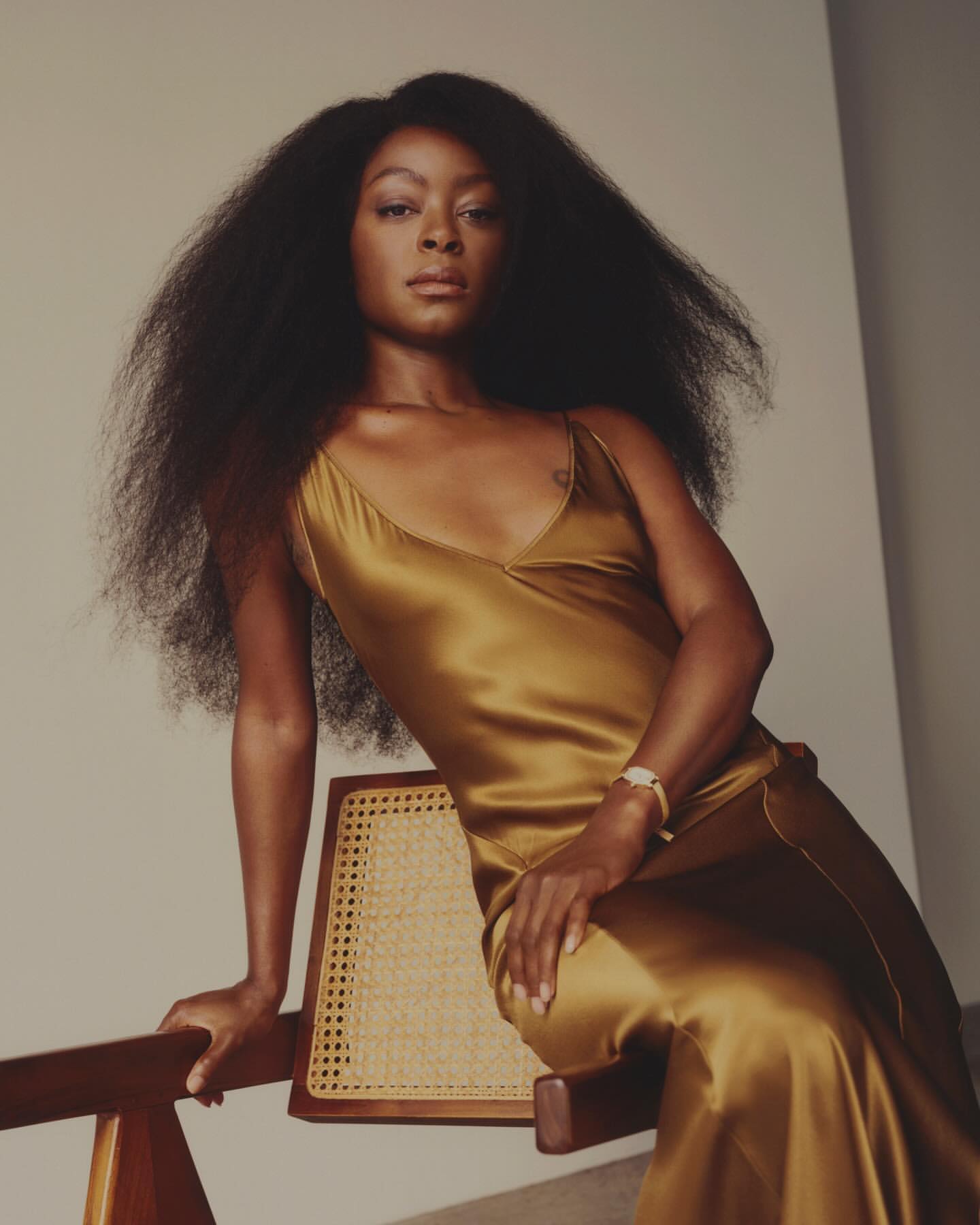 Black woman with long voluminous hair wearing a gold silk slip dress, seated on a wooden chair against a plain wall.