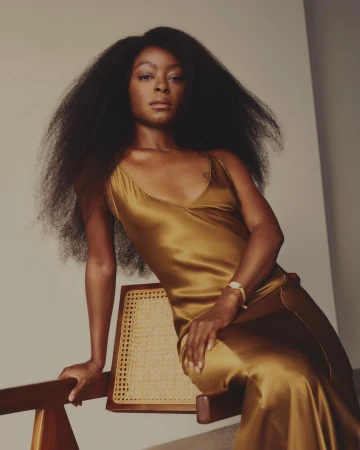 Black woman with long voluminous hair wearing a gold silk slip dress, seated on a wooden chair against a plain wall.