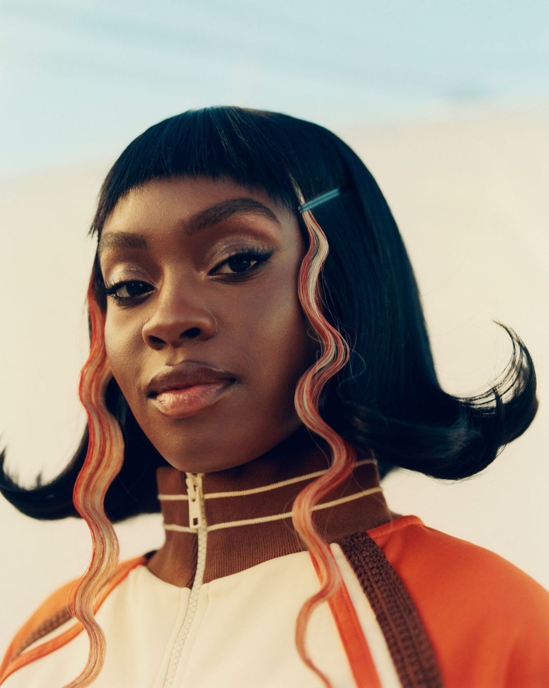 Close-up portrait of an adult Black female model with black hair, light skin, dark eyes, wearing a white and orange jacket, indoors.