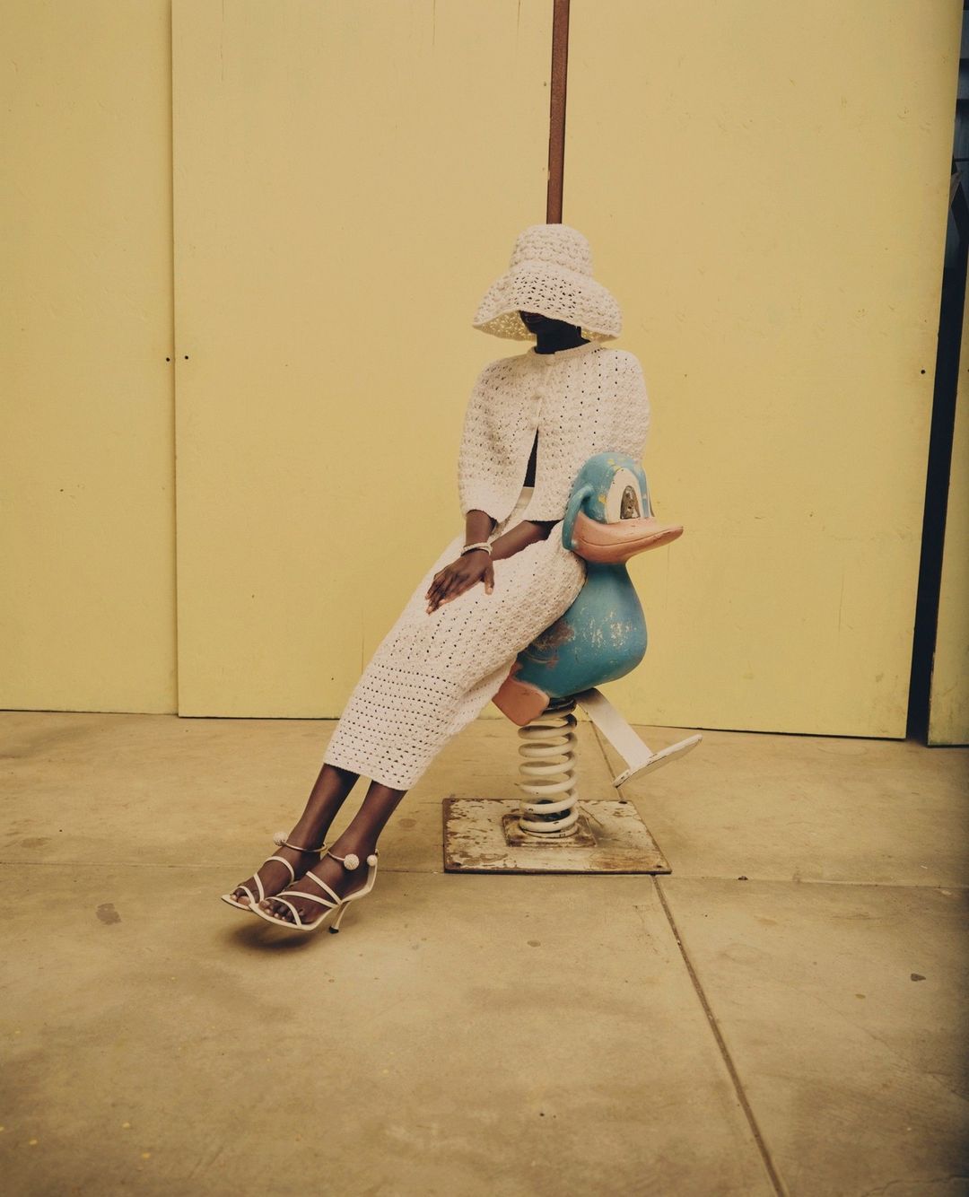 Woman in coordinated crochet hat, jacket, and skirt sits on a blue duck-shaped spring rider against a yellow wall in bright daylight.