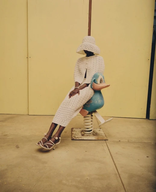 Woman in coordinated crochet hat, jacket, and skirt sits on a blue duck-shaped spring rider against a yellow wall in bright daylight.