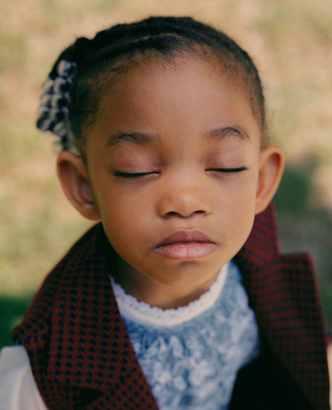 Portrait of an African-American child with braided hair and a red vest, eyes closed, in a serene outdoor setting.