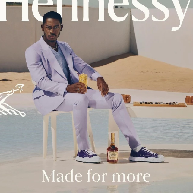 A Black male model in a lilac suit is seated with a cocktail. The scene includes Hennessy bottle, a sandy background, and branding.