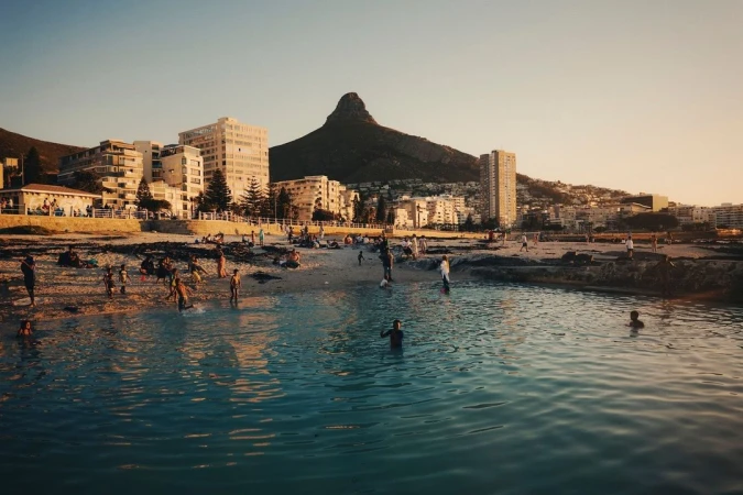 People swimming and gathering at a coastal tidal pool at sunset with buildings and a prominent mountain peak in the background.