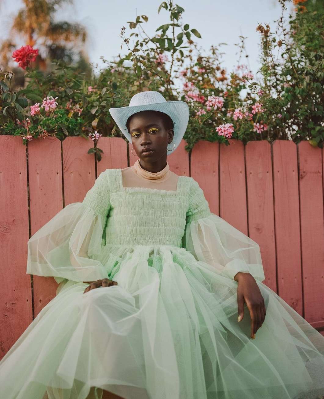 Woman in a pastel green dress and cowboy hat sits in front of a pink fence and flowering bushes, gazing calmly into the camera.