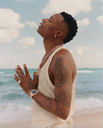 Male celebrity in a sleeveless top poses on a beach, facing the sky. He wears jewelry and displays tattoos on his arms.
