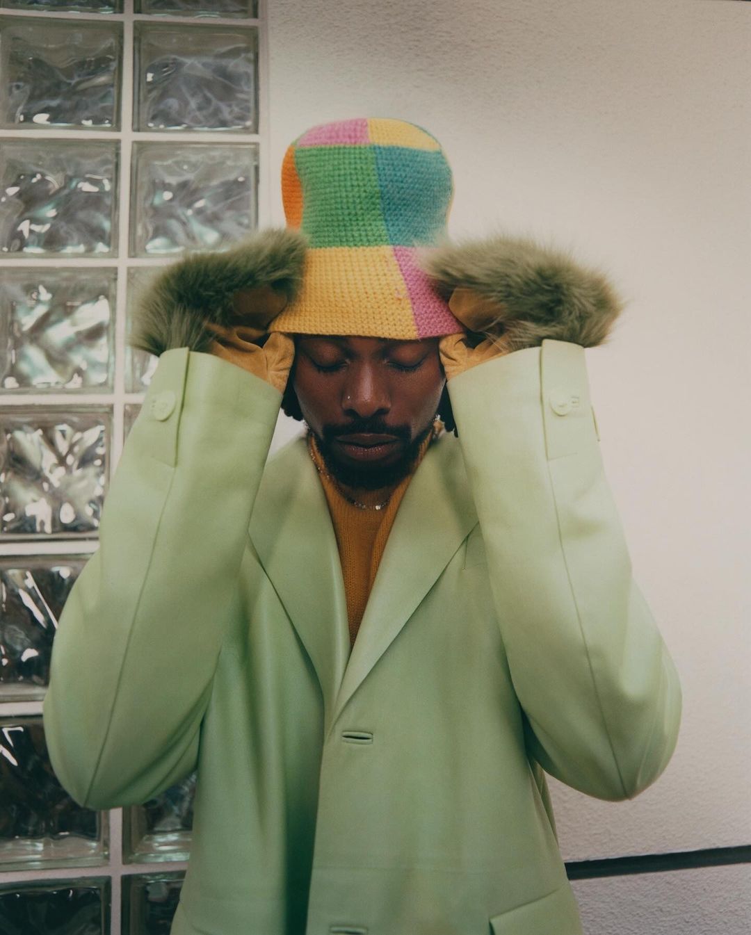 Portrait of an adult male wearing a bucket hat and light green jacket with hands near his face, standing indoors near a glass block wall.