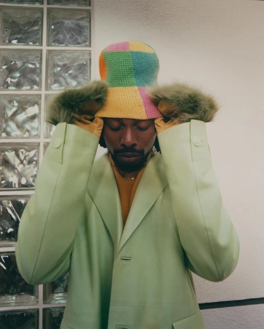 Portrait of an adult male wearing a bucket hat and light green jacket with hands near his face, standing indoors near a glass block wall.