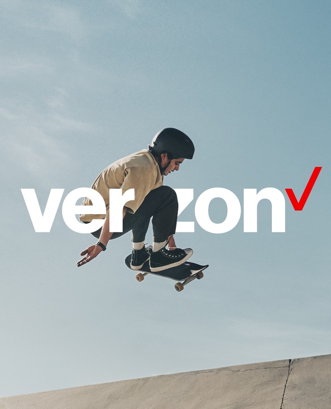 A young adult man in mid-air on a skateboard, wearing a helmet and casual clothes, against a bright sky with the Verizon logo.