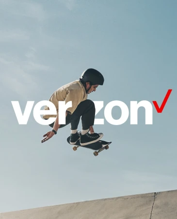 A young adult man in mid-air on a skateboard, wearing a helmet and casual clothes, against a bright sky with the Verizon logo.