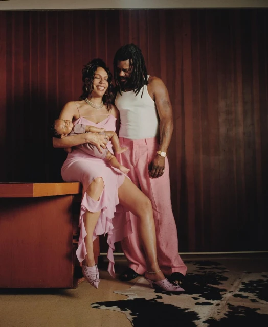 Family portrait featuring a smiling woman in a pink dress holding a baby, with a bearded man in a white tank top in the background.