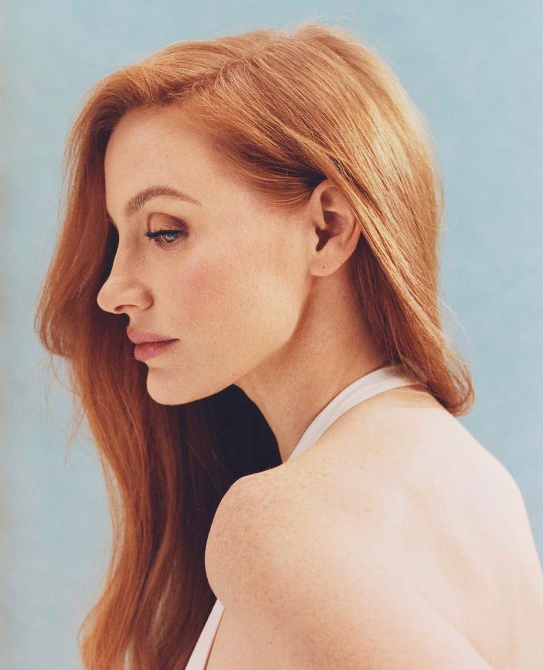 Profile portrait of a fair-skinned woman with long red hair wearing a white top against a light blue background.