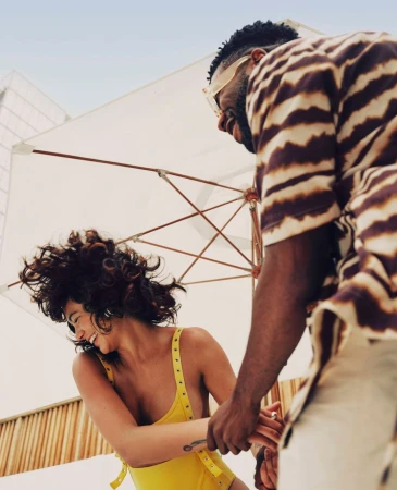 A smiling woman in a yellow swimsuit and a man in a patterned shirt standing outdoors under a large white sun umbrella.
