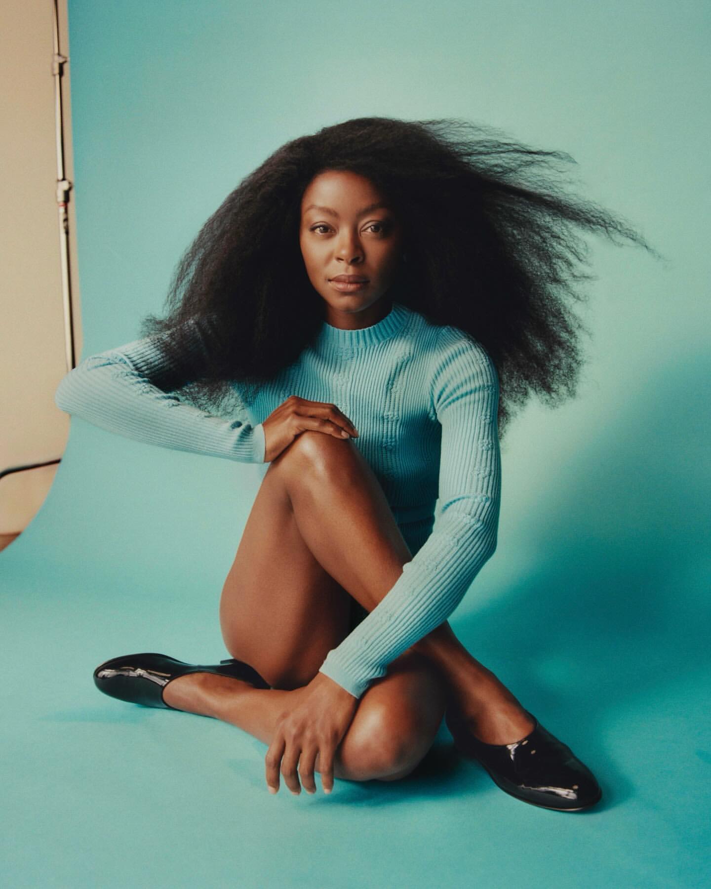 Portrait of an adult woman with afro textured hair wearing a light blue long sleeve shirt and black shoes sitting on a blue surface.
