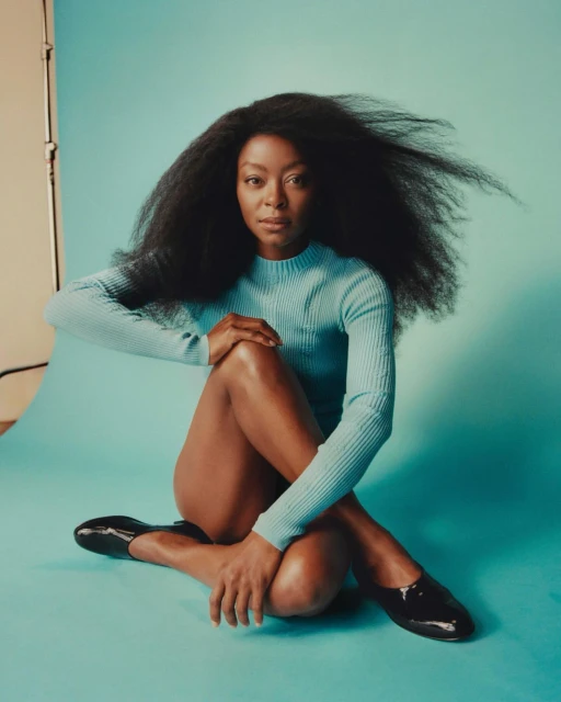 Portrait of an adult woman with afro textured hair wearing a light blue long sleeve shirt and black shoes sitting on a blue surface.