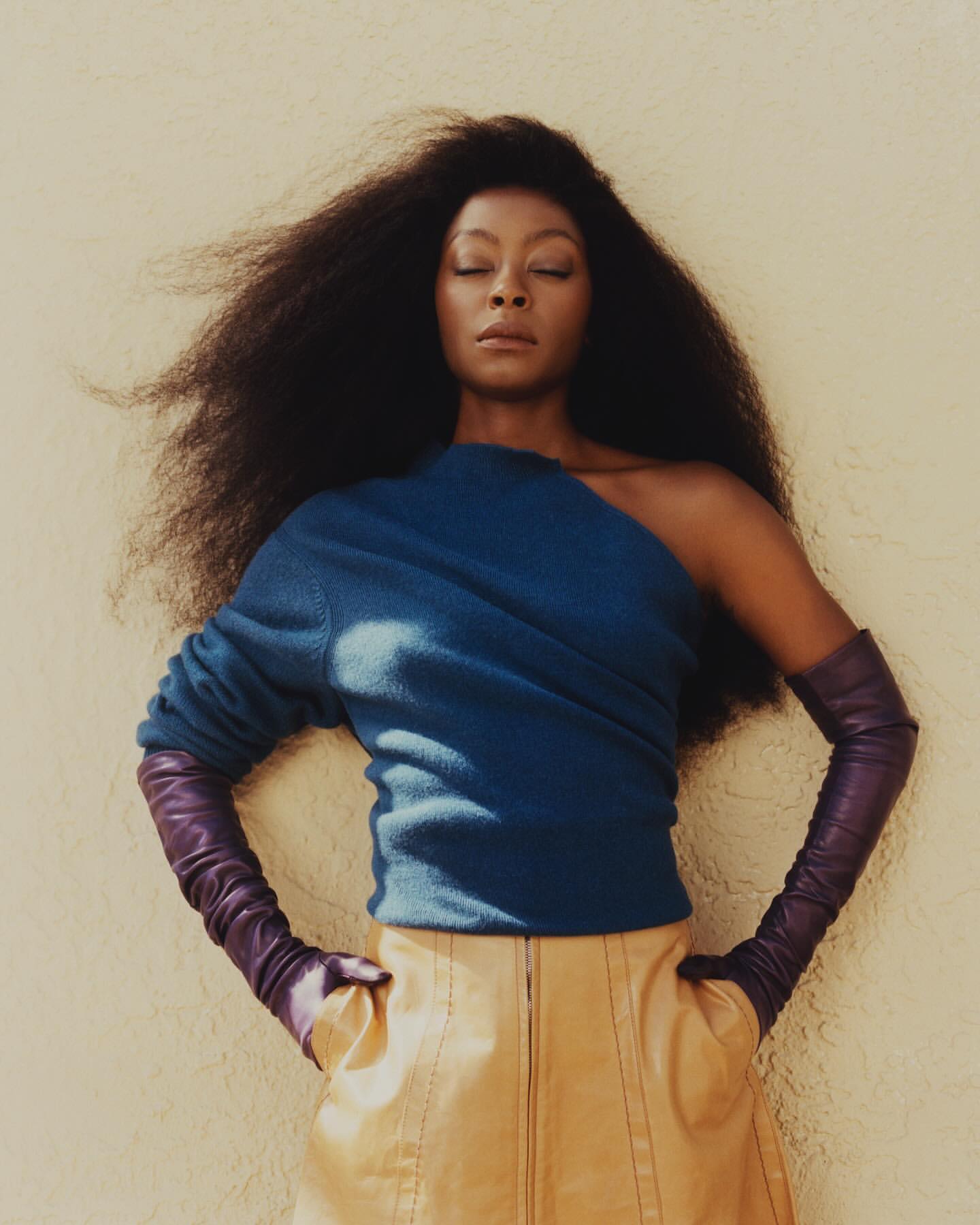 A woman with an afro poses confidently in a studio, wearing a blue top, leather skirt, and gloves, lit by soft light.