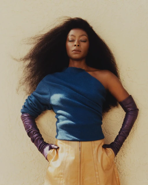A woman with an afro poses confidently in a studio, wearing a blue top, leather skirt, and gloves, lit by soft light.