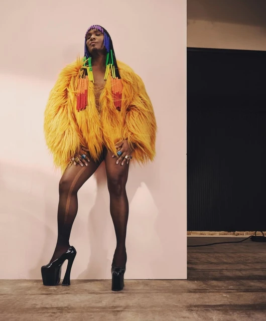 Person poses in bright orange faux-fur coat with rainbow beaded headdress, sheer black hose, and platform heels against pink wall.