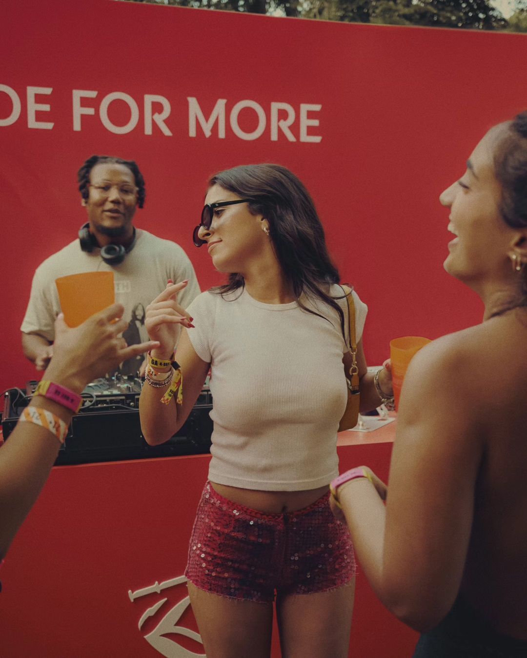 Two women in casual summer outfits laugh at an outdoor event, with a DJ in the background and a red promotional backdrop.