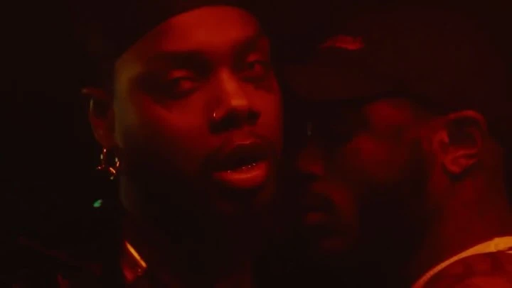 Two men in close proximity in a dark, moody setting with red lighting, wearing caps and earrings.