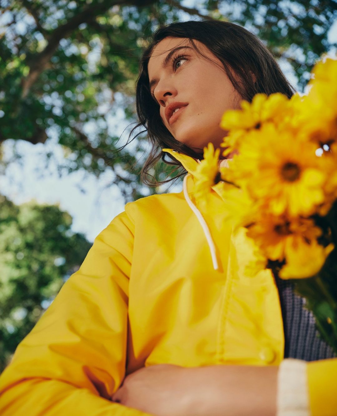 A young adult woman wearing a yellow jacket poses with yellow flowers outdoors against a background of green trees and blue sky.
