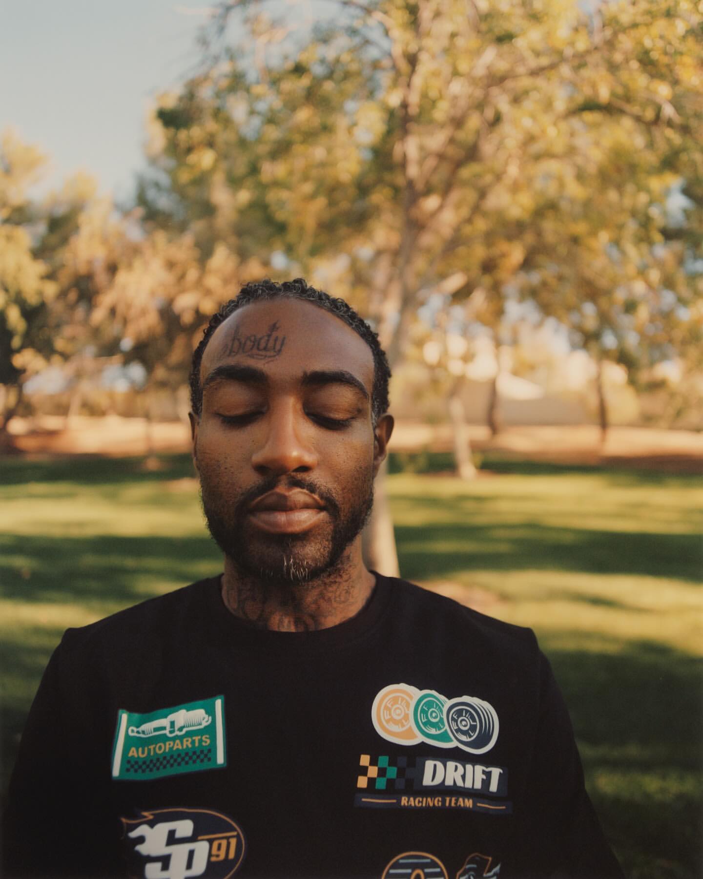 Adult man with eyes closed wearing a black shirt with race car patches stands in an outdoor park setting with trees in the background.