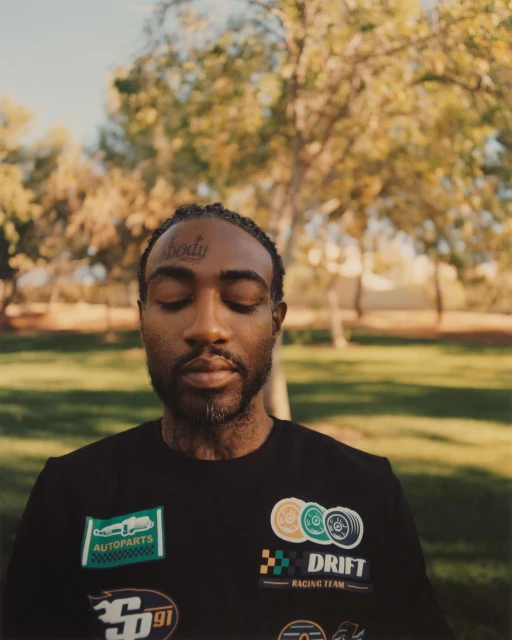 Adult man with eyes closed wearing a black shirt with race car patches stands in an outdoor park setting with trees in the background.