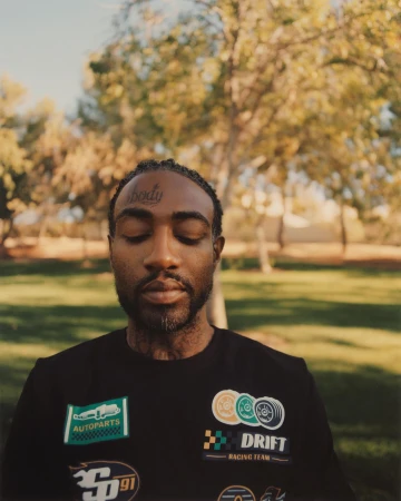 Adult man with eyes closed wearing a black shirt with race car patches stands in an outdoor park setting with trees in the background.