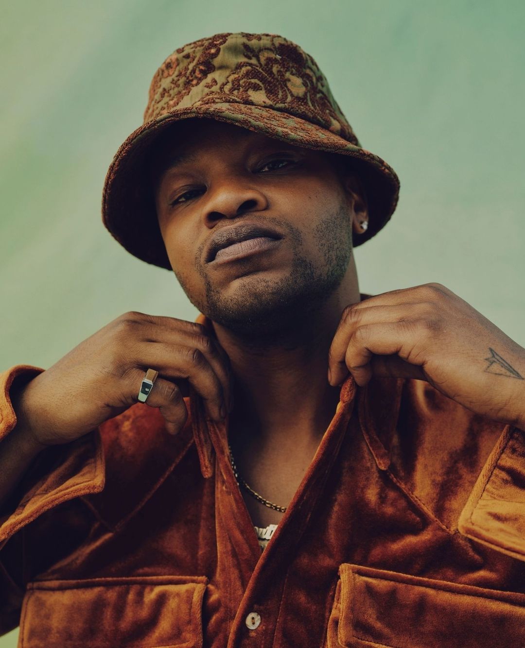 Portrait of an adult male wearing an ornate bucket hat and velvet shirt, set against a muted green backdrop, with a medium studio shot.