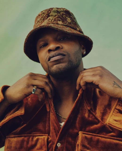 Portrait of an adult male wearing an ornate bucket hat and velvet shirt, set against a muted green backdrop, with a medium studio shot.