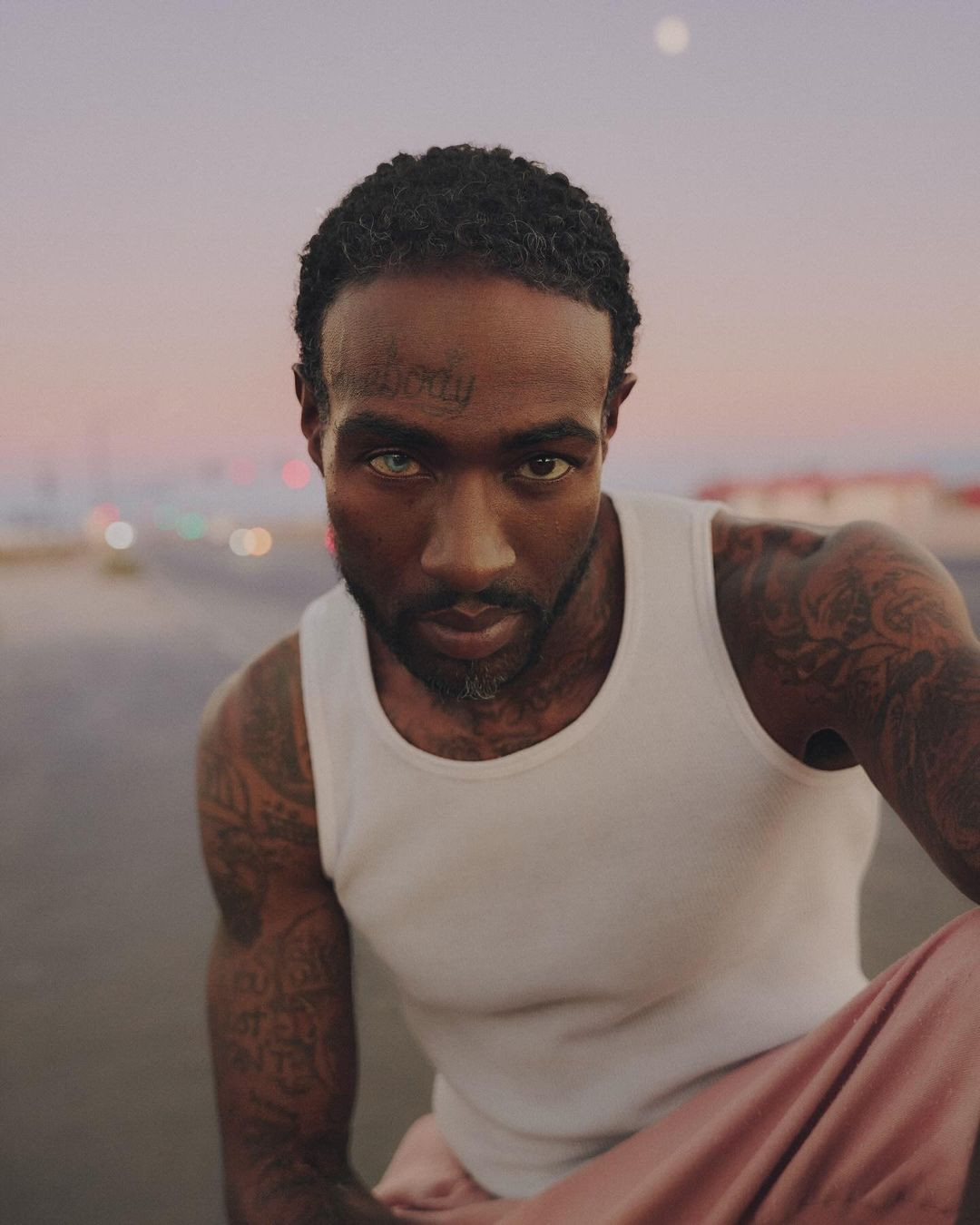 Portrait of a black man with tattoos and a white tank top, gazing intensely at the camera against a muted urban backdrop.