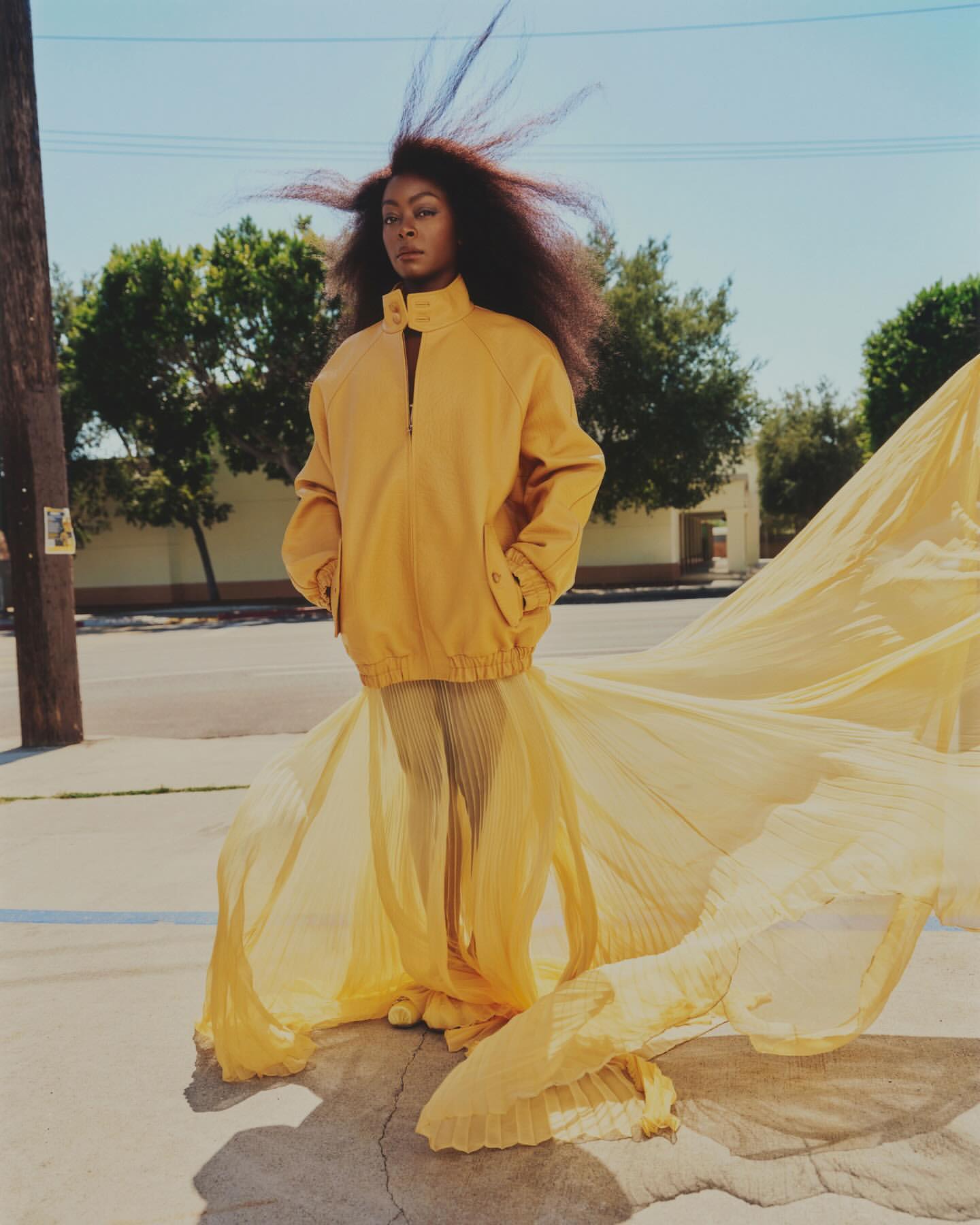 A person in a yellow jacket and skirt standing on a city street. The skirt has a long flowing train billowing out behind her.