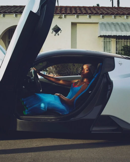 A fashionable Black woman in a blue dress sits inside a luxury sports car with the door open, posing on a sunny street outside a house.