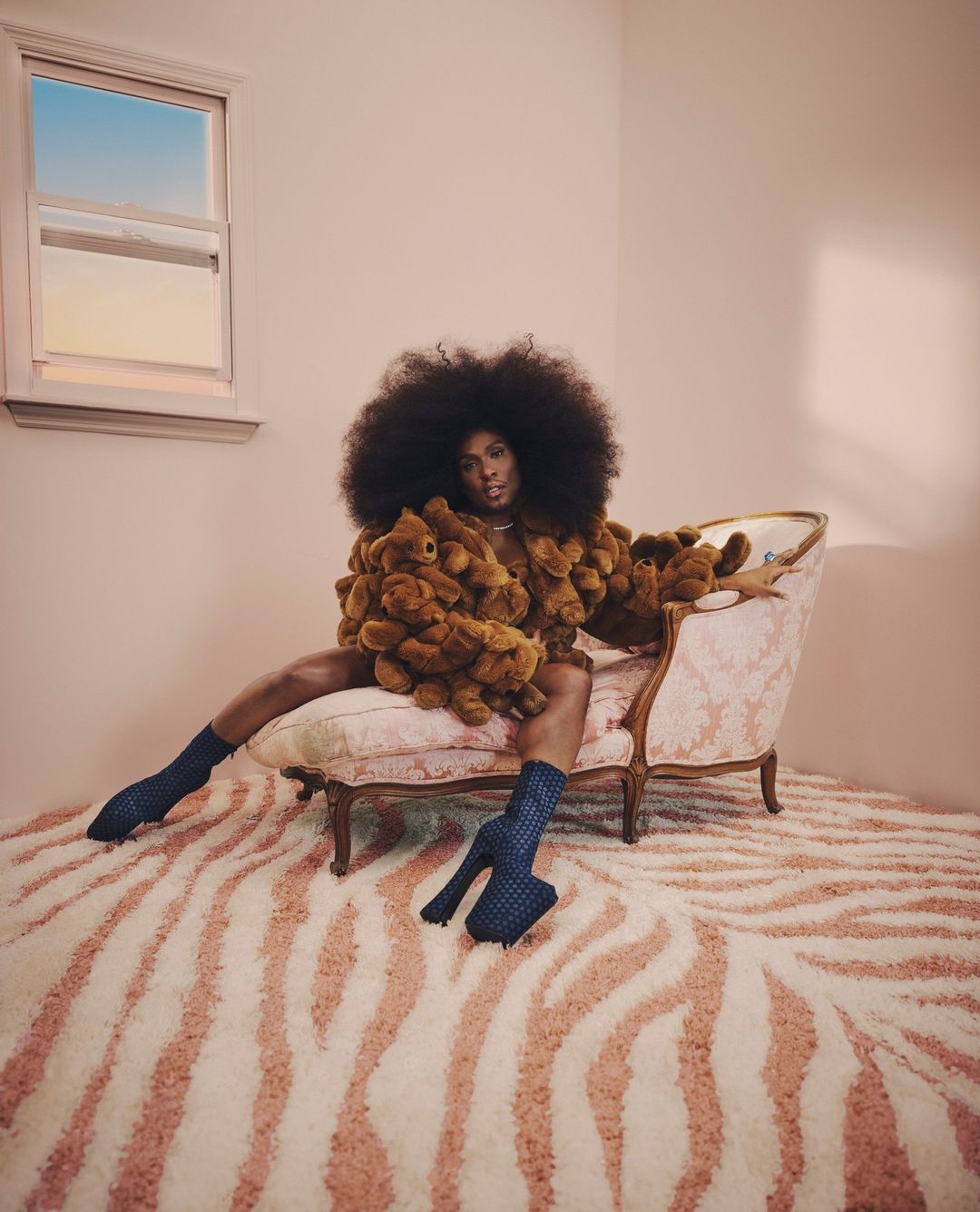 A Black model poses in a room with a window, seated on an antique chair and wearing a jacket made of teddy bears and blue heeled socks.