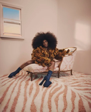 A Black model poses in a room with a window, seated on an antique chair and wearing a jacket made of teddy bears and blue heeled socks.