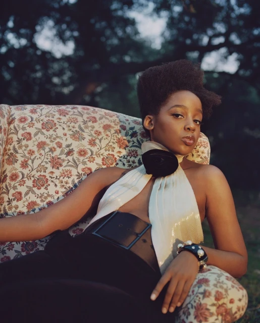 A young Black woman lounges on a floral armchair outdoors, wearing a cream halter top, black pants, and a pearl bracelet.