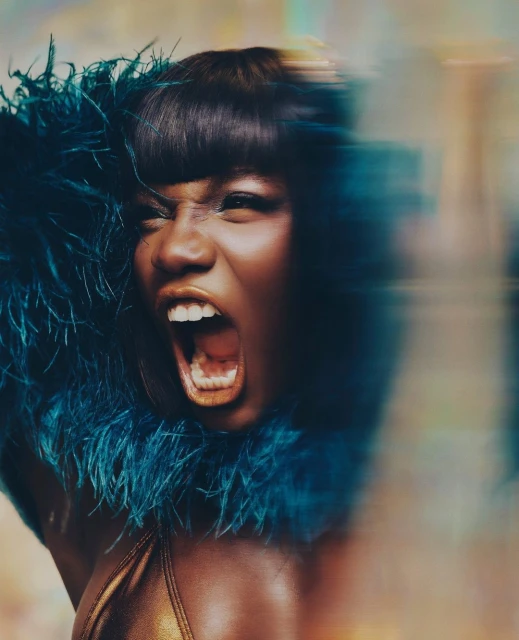 Close-up portrait of an adult woman with dark hair and blue feather boa yelling with mouth open showing teeth in a brightly lit scene.